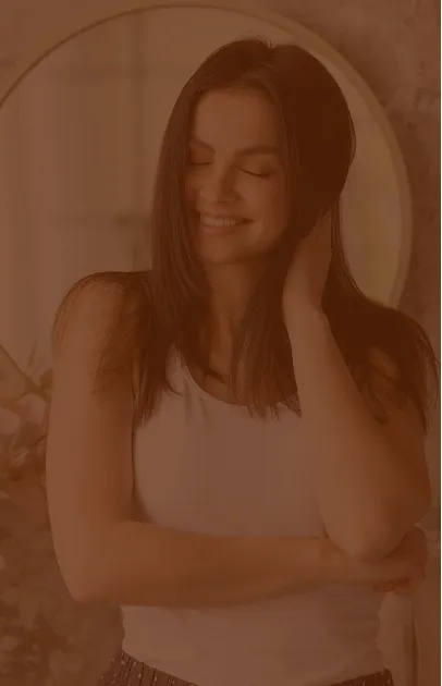 Smiling woman with long brown hair standing in front of a round mirror, wearing a white sleeveless top.