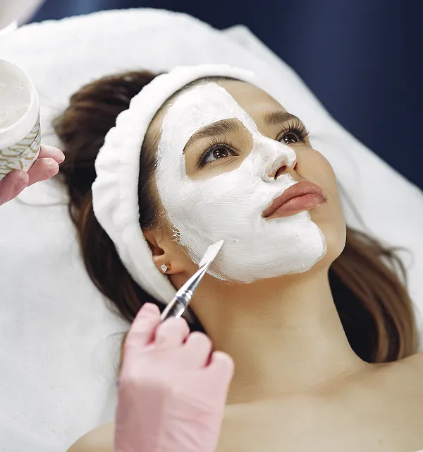 Woman lying down with a white facial mask being applied by a gloved hand using a brush.