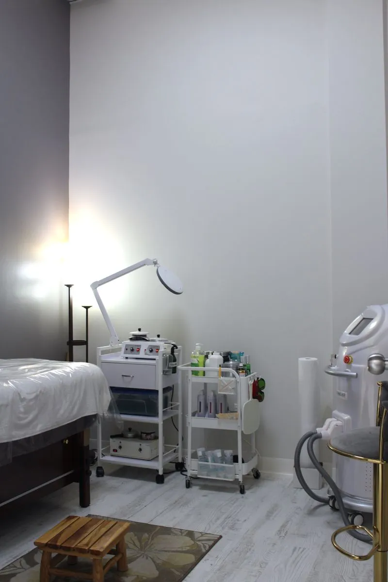 Clean and organized spa treatment room with a bed covered in plastic, skincare equipment, a small wooden stool, and a floor lamp.