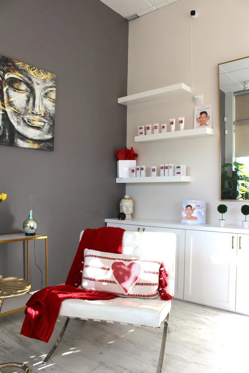 Bright stylish reception area with white chair draped in red blanket and heart-patterned pillow, white shelves with skincare products, and Buddha artwork.