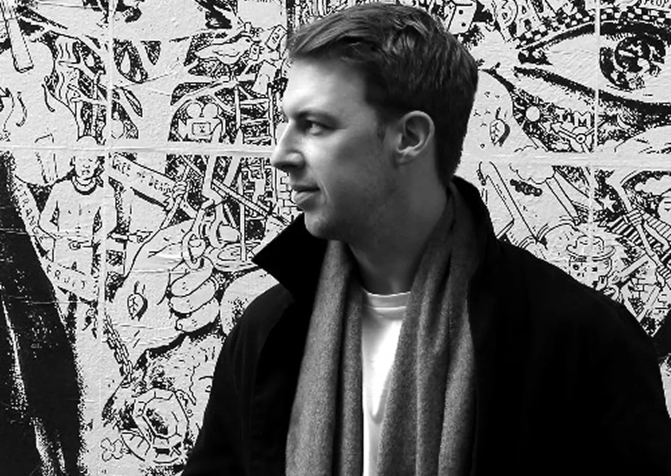 Black and white photo of Jamie Neasham in a coat and scarf looking to the side in front of a detailed mural wall in Paris, France.