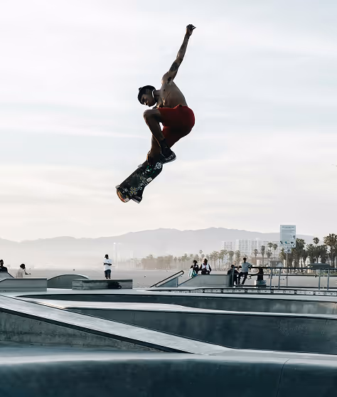 Shirtless skateboarder in red shorts performing a high jump trick at a skate park with mountains and palm trees in the background.