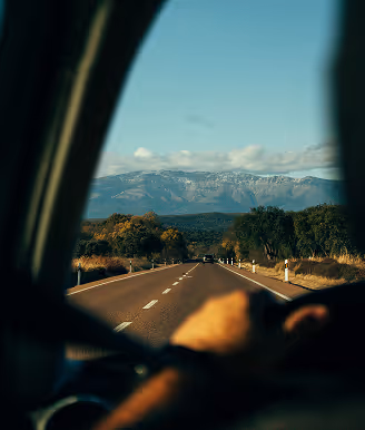 View of a straight road leading towards mountains, seen from inside a car with a driver's hand on the steering wheel.