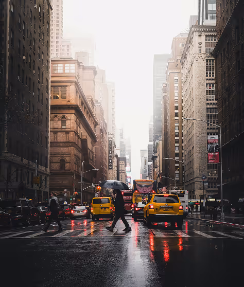 People crossing a wet city street with yellow taxis and tall buildings on a cloudy day.