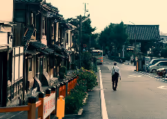 Person walking alone down a quiet street lined with traditional-style buildings and parked cars in the distance.