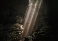 Sunlight beams shining through an opening into a dark cave with rocky walls.
