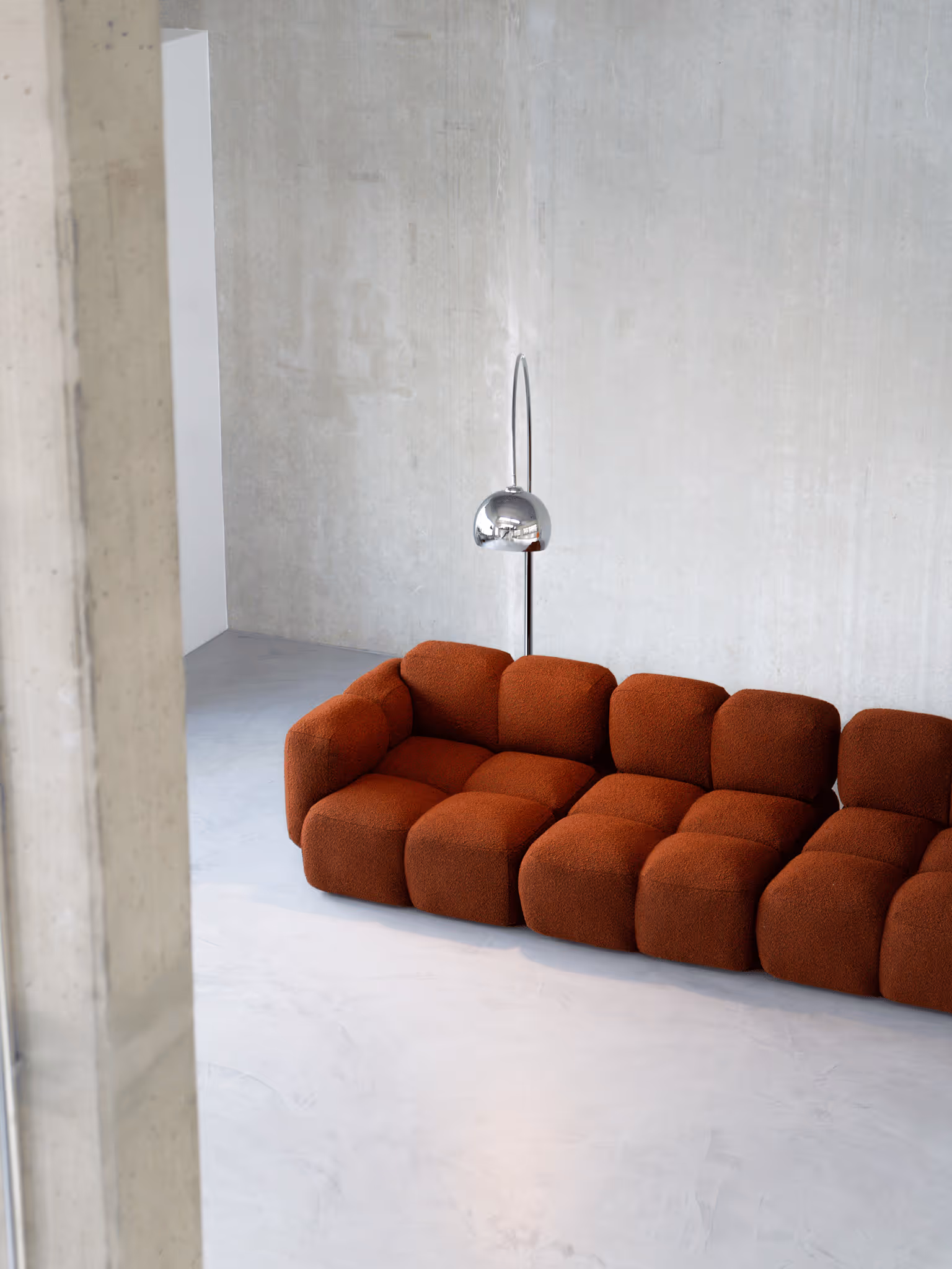 Seitliche Perspektive auf rostbraunes modulares Bouclé-Sofa mit verchromter Bogenlampe in Betonraumecke