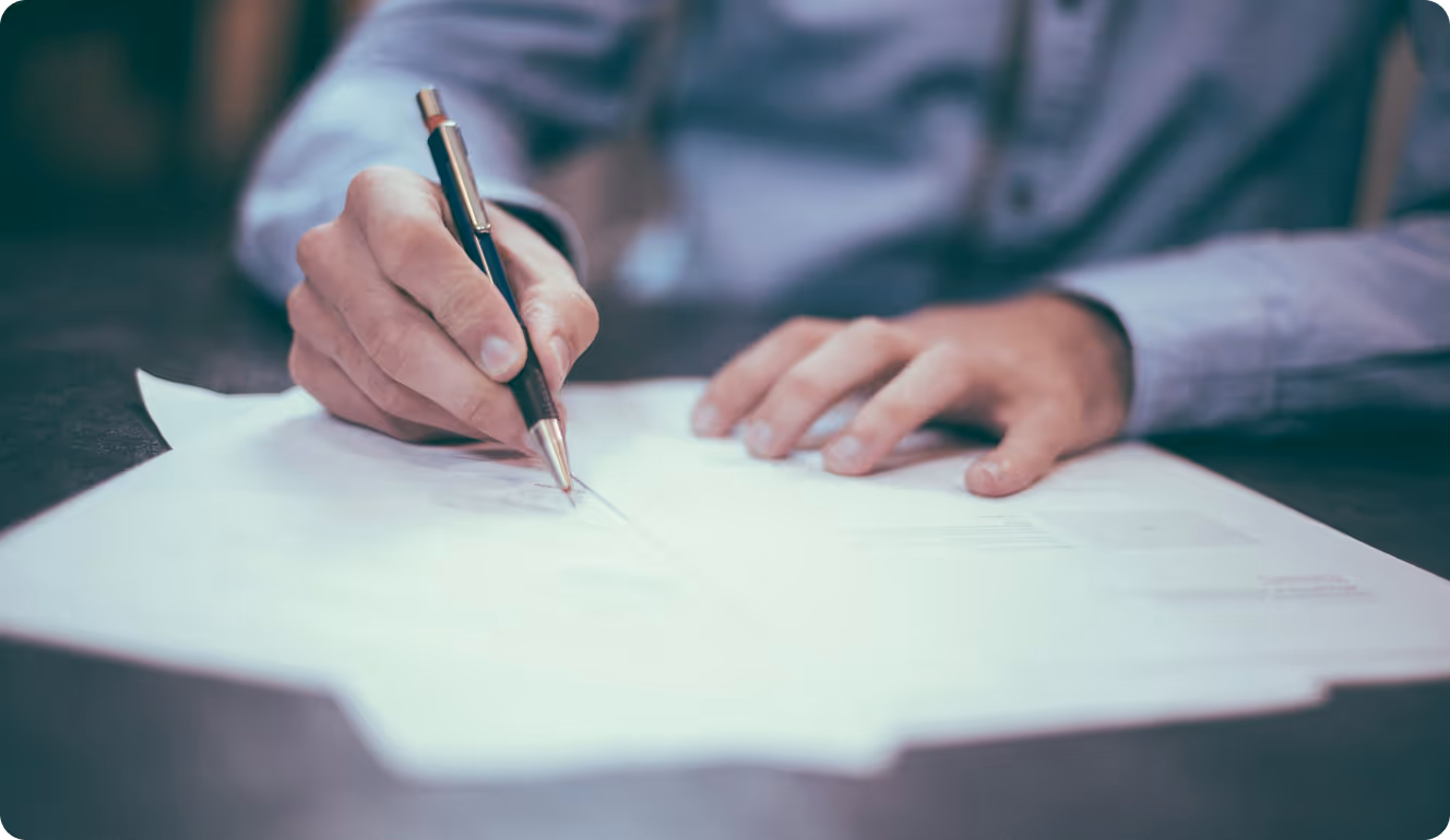 Person signing or writing on a document with a pen on a desk.