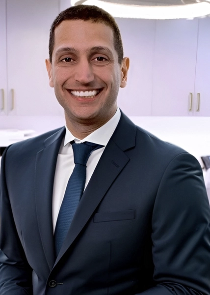 Smiling man in a dark suit, white shirt, and blue tie standing in a modern office kitchen.