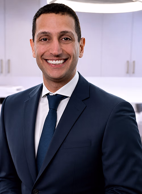 Smiling man in a dark suit, white shirt, and blue tie standing in a modern office kitchen.