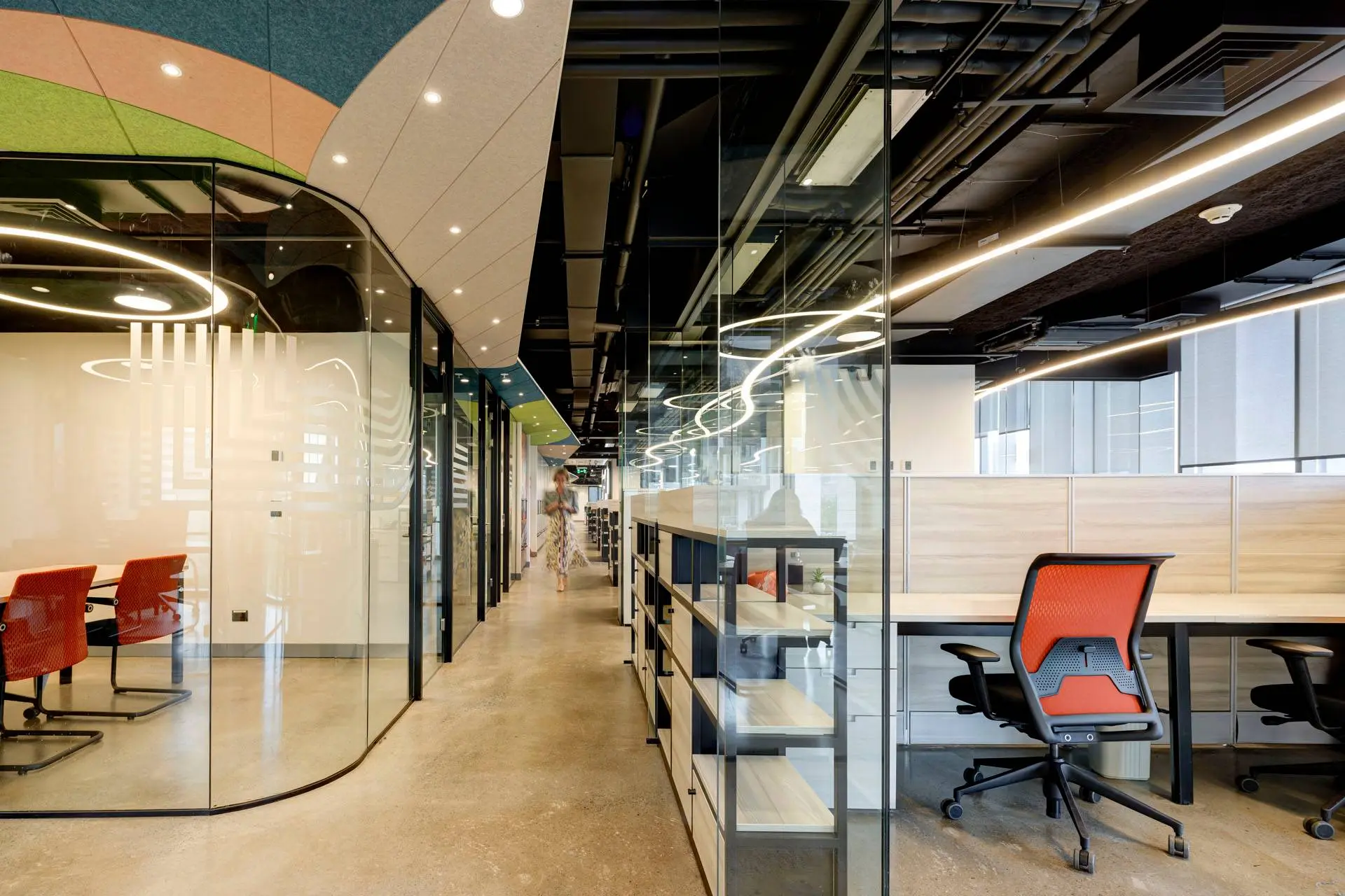 Oficina moderna con pasillo central, escritorios con separadores y plantas decorativas, y una persona caminando.