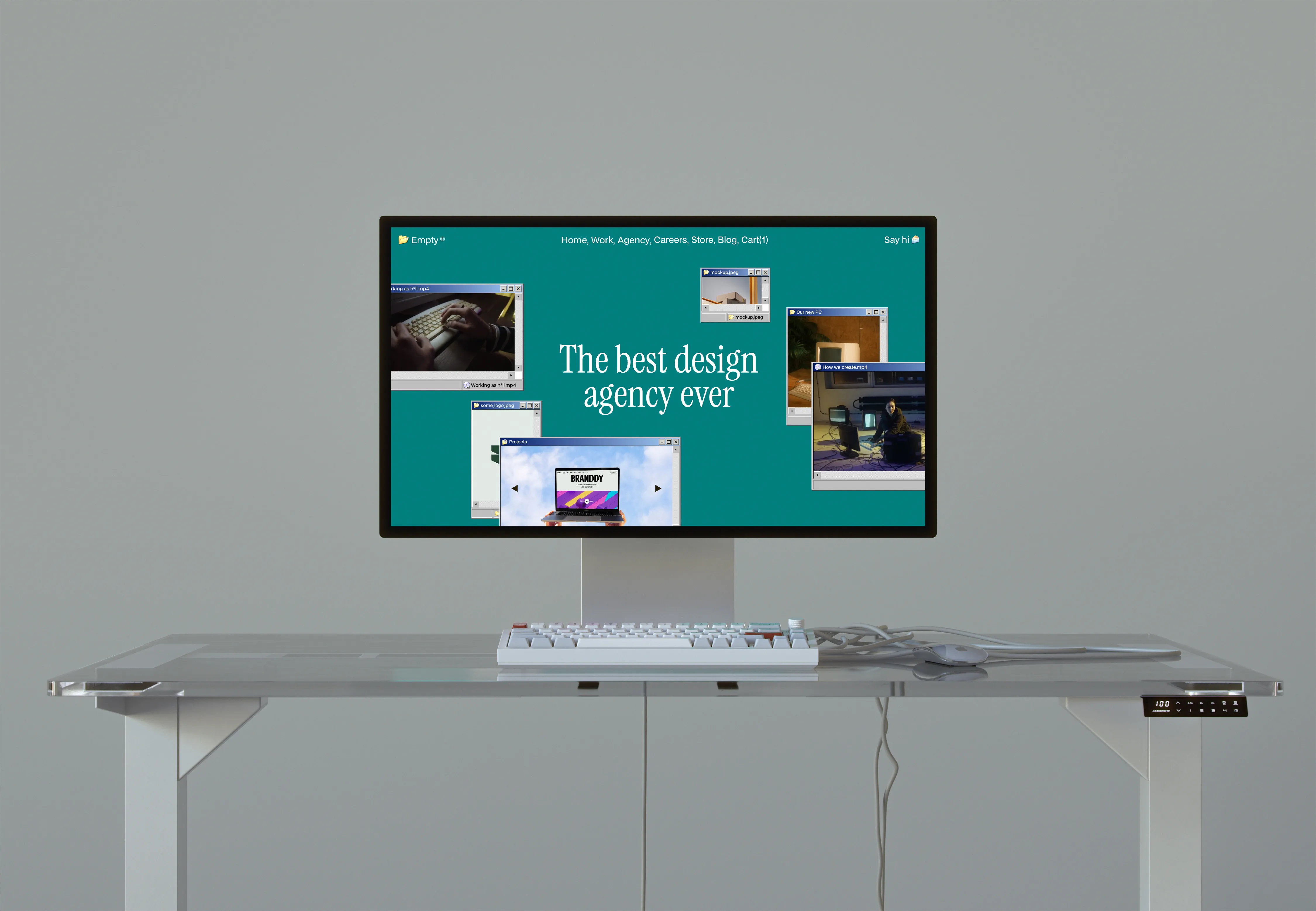 Minimalistic workspace with a transparent glass desk, white mechanical keyboard, mouse, and a monitor displaying a website homepage reading 'The best design agency ever'.
