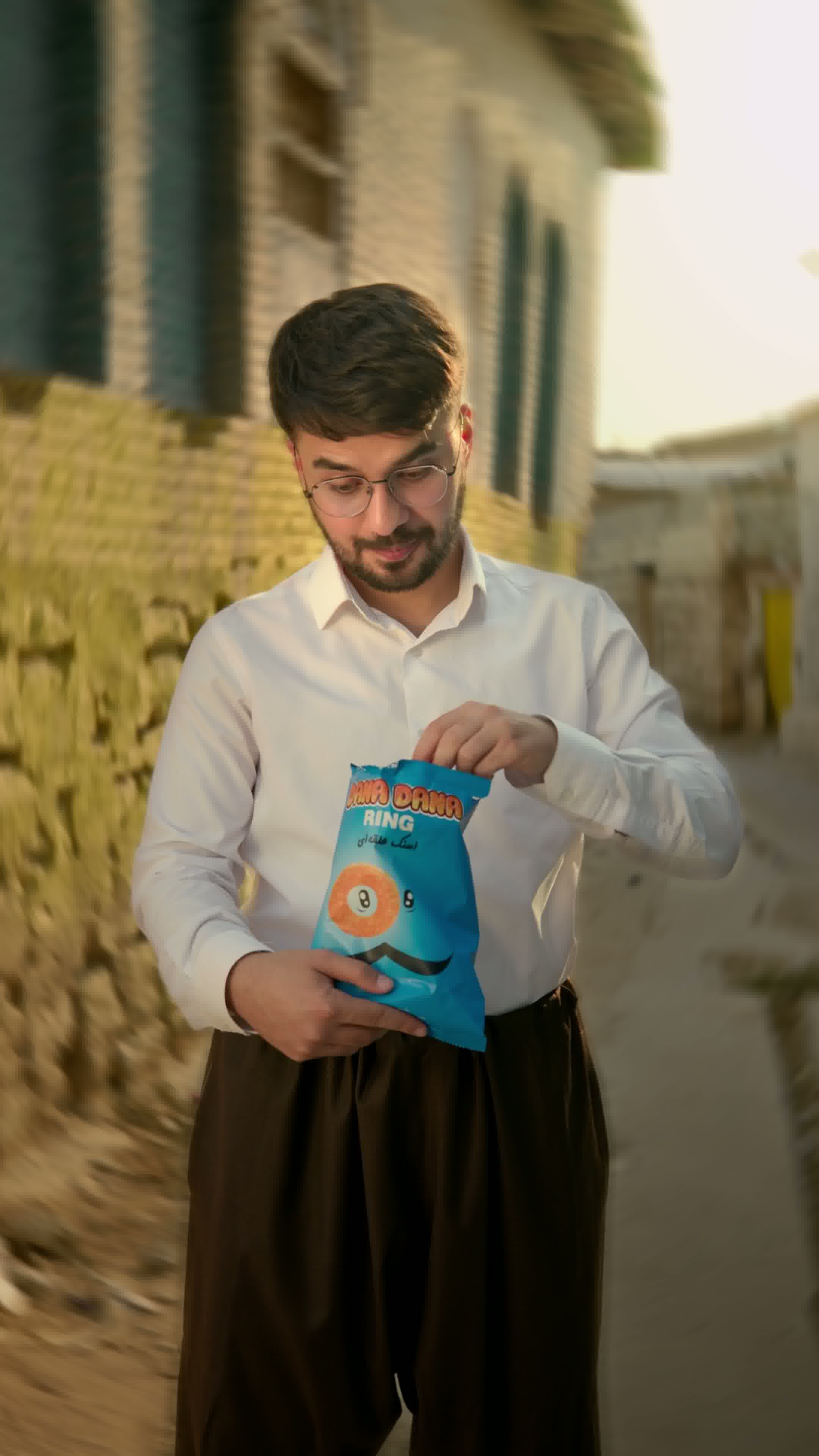 Young man with glasses wearing a white shirt and dark pants opening a blue snack bag outdoors in a sunlit alley.