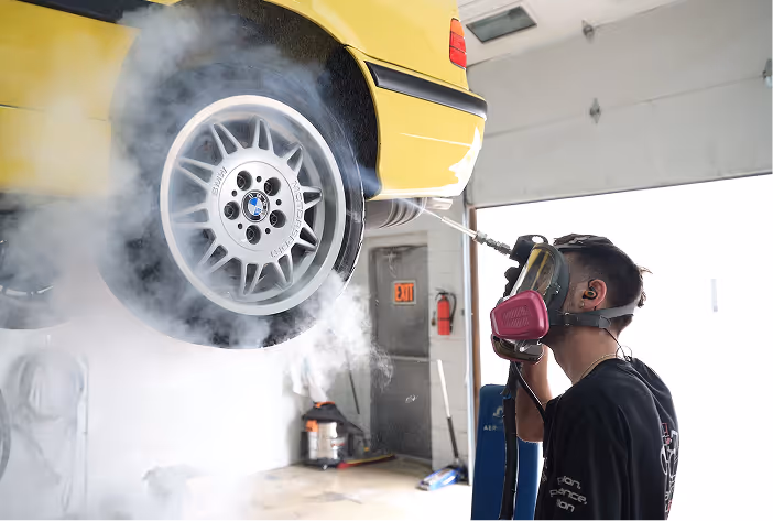 Person wearing a respirator mask using a pressure washer to clean the wheel of a yellow car in a garage.
