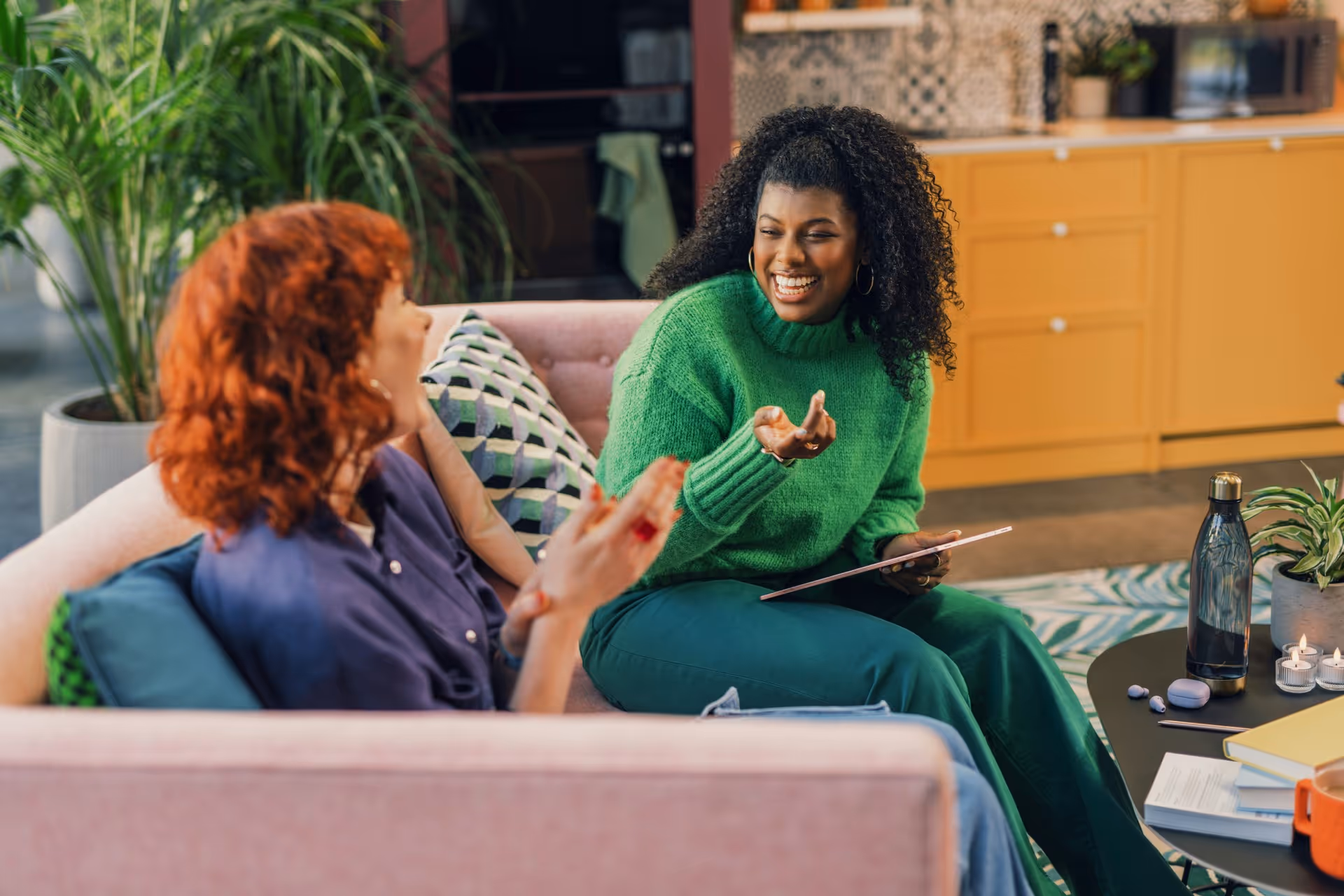 Two women sit on a sofa in a cozy room, engaged in a lively conversation. One wears a green sweater, the other a blue shirt. Both are smiling warmly.