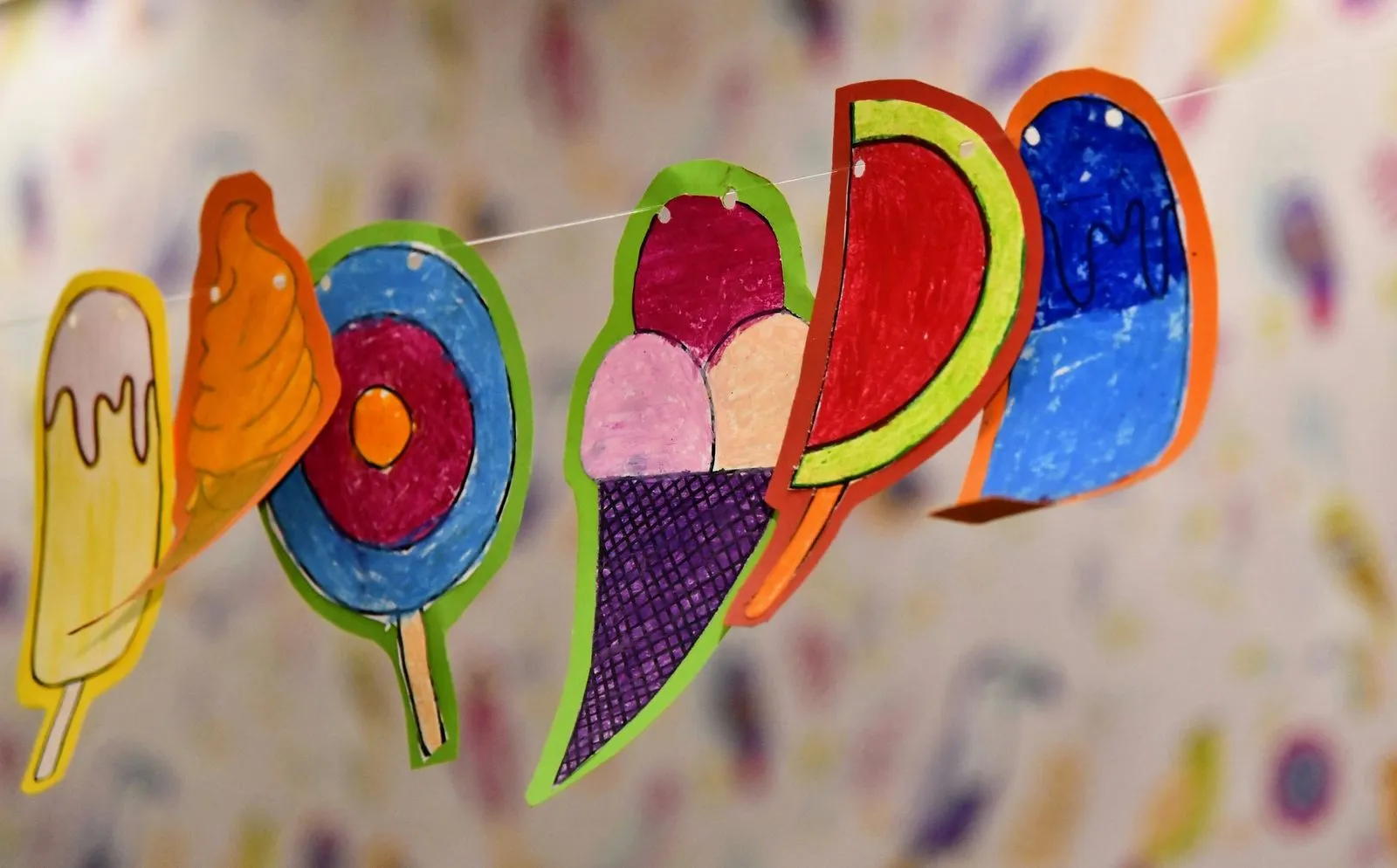 Colorful paper drawings of various ice creams and popsicles hanging on a string, set against a blurred, playful background, evoking a joyful, whimsical mood.