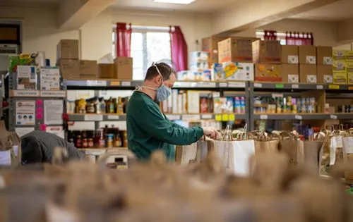 A man wearing a mask and green sweater packs paper bags in a food pantry, surrounded by shelves of canned goods and boxes. The mood is focused and industrious.