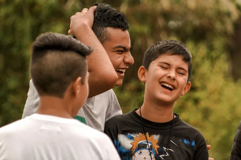 Three boys outdoors, smiling and laughing together. One boy playfully touches another's head. Their joyful expressions convey happiness and friendship.