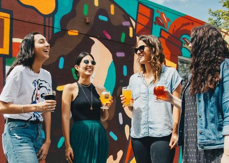 Four people laughing and holding drinks in front of a colorful mural. The vibrant background and their smiles convey a joyful, lively atmosphere.