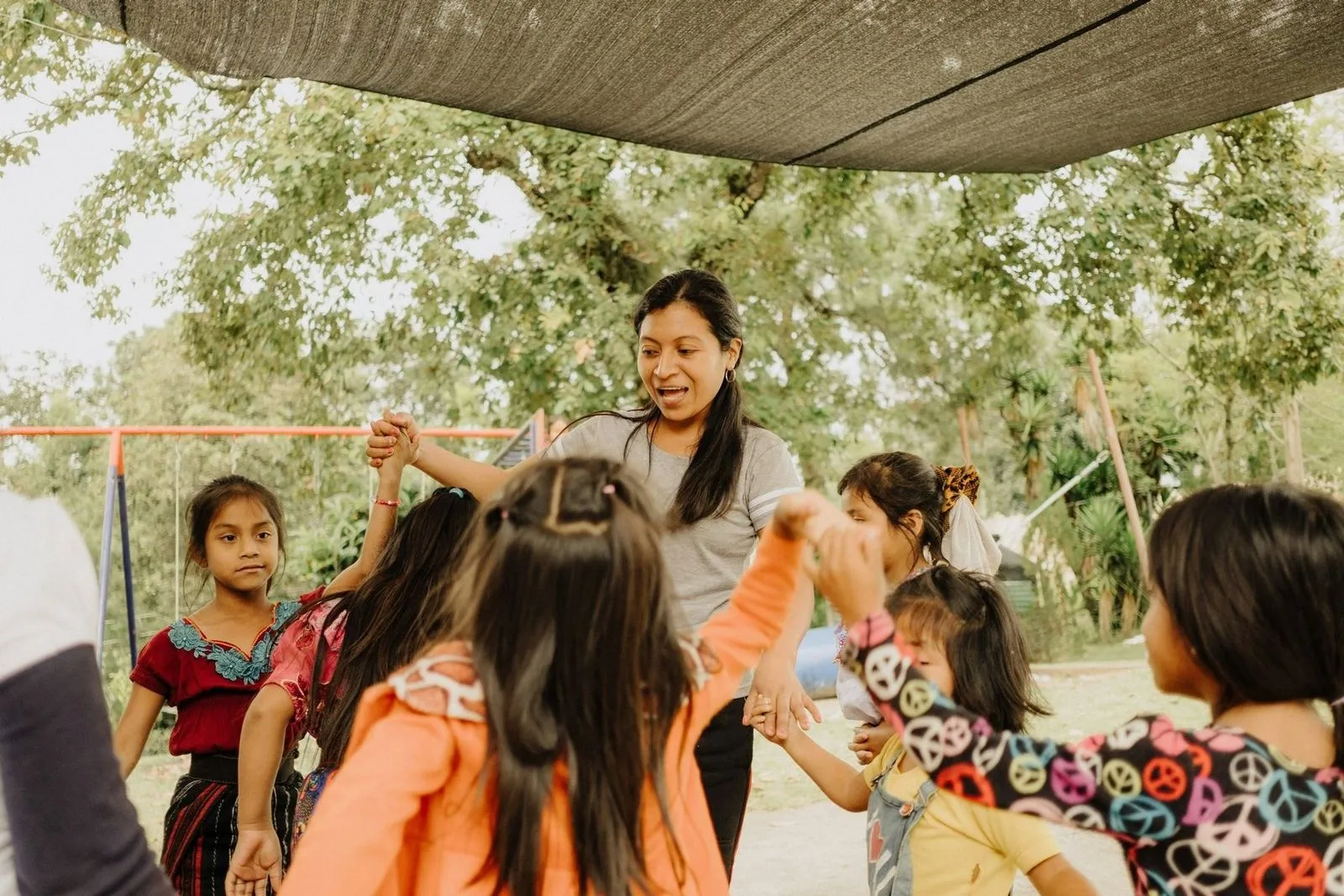 A woman and several children hold hands in a circle outdoors, under a canopy. The children wear colorful clothing, and their expressions convey joy and engagement. Trees and playground equipment are visible in the background.