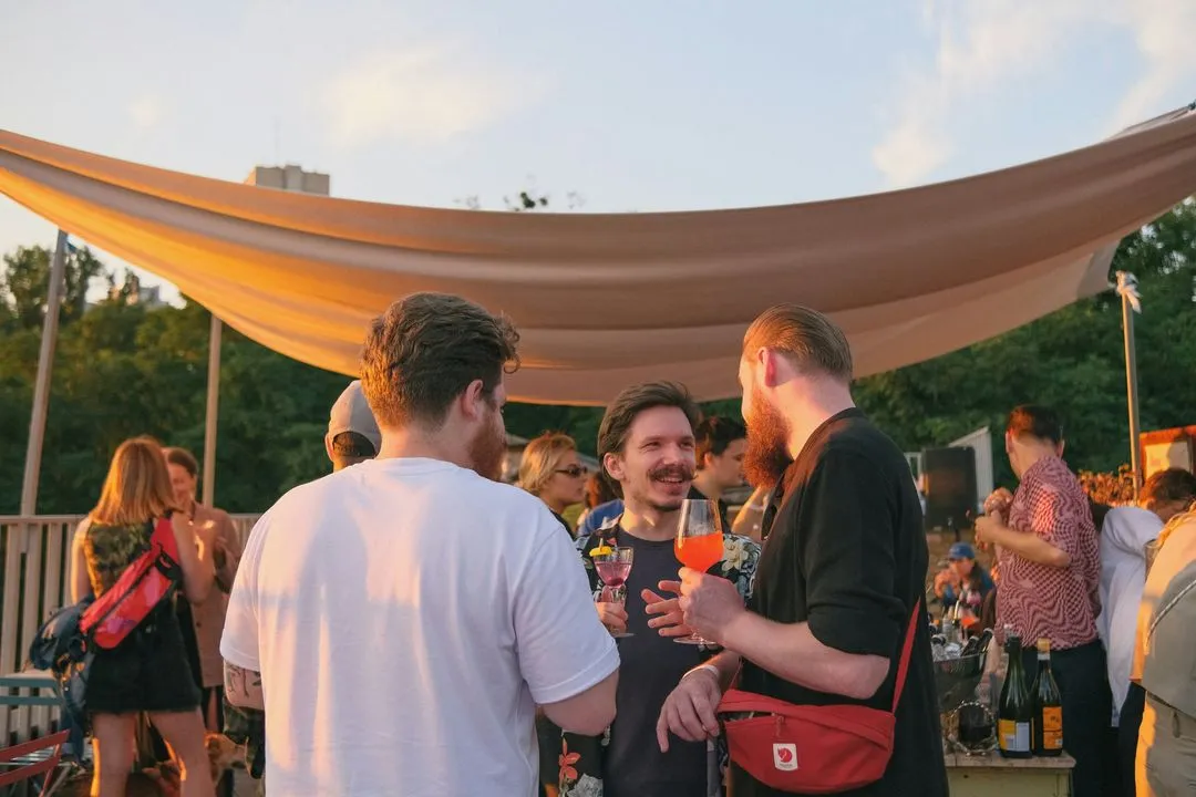 A group of people socializing at an outdoor party during sunset. They hold colorful drinks, smiling and chatting under a canopy, creating a lively and warm atmosphere.