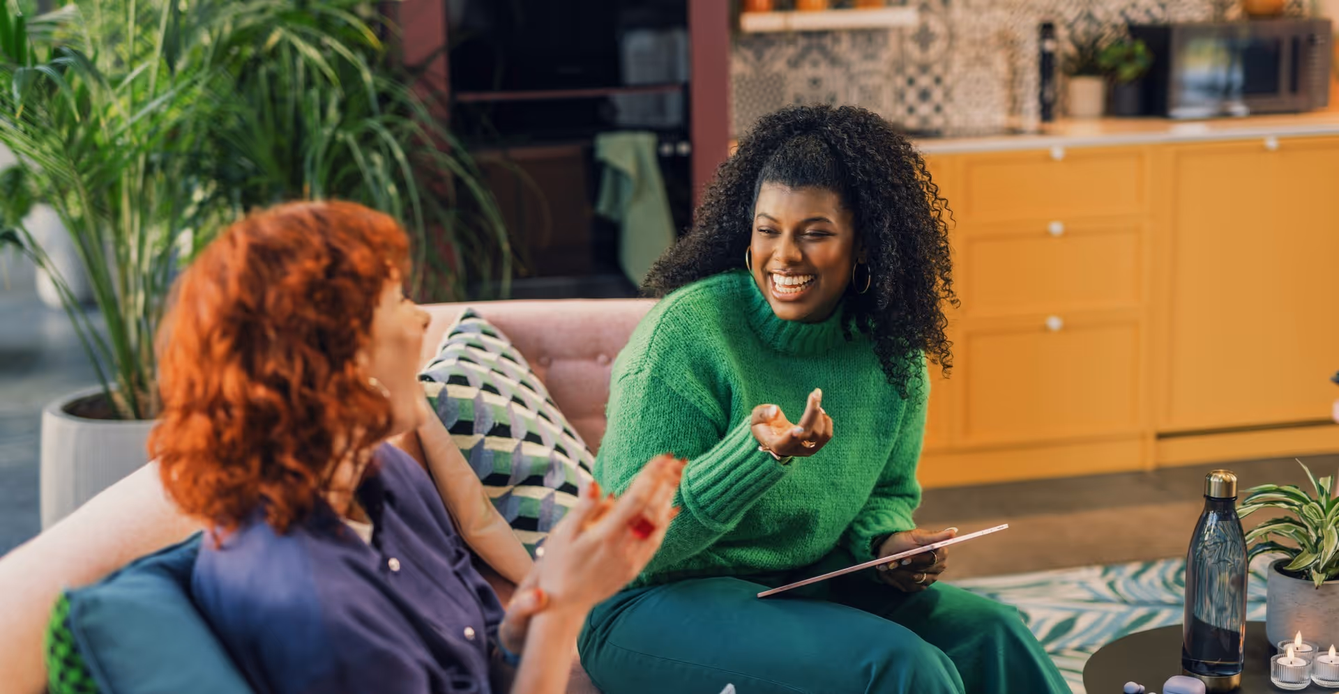 Two women sit on a sofa, laughing and chatting. One wears a green sweater, holding a tablet. The room is cozy with plants and yellow cabinets.
