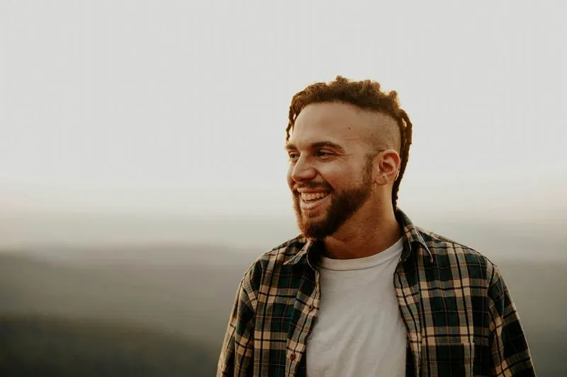 A man with a beard and styled hair smiles warmly while looking to the side. He's wearing a plaid shirt over a white t-shirt against a blurred scenic background.