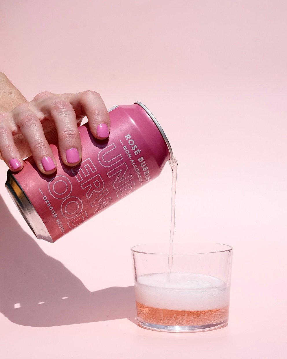 Hand with pink-painted nails pouring Underwood NA Rosé Bubbles from a pink can into a clear glass on a pink background.