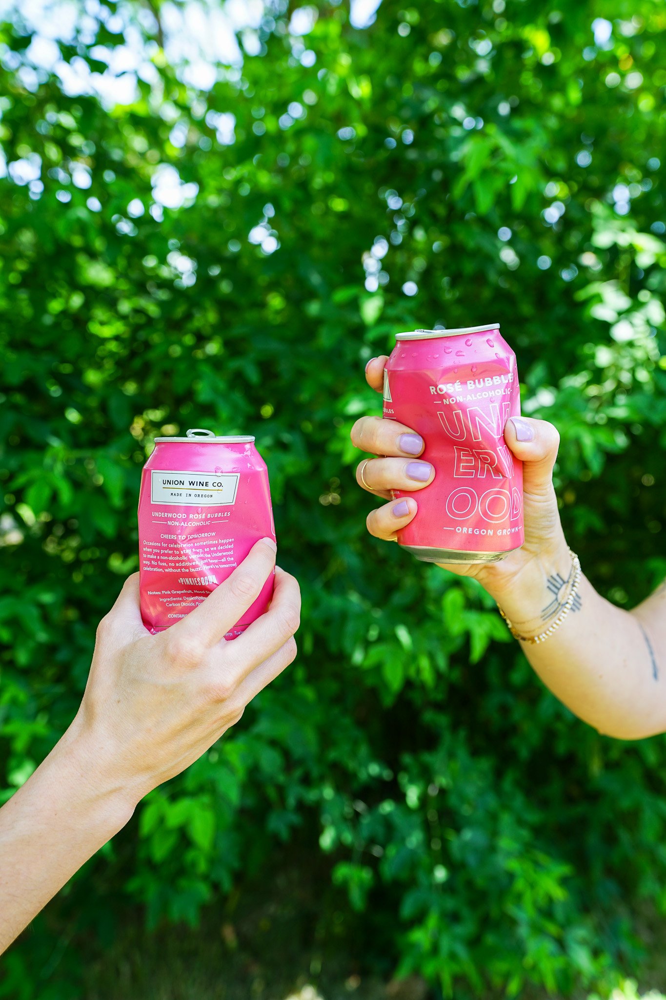 Two hands holding pink cans of Underwood NA Rosé Bubbles against a green leafy background.