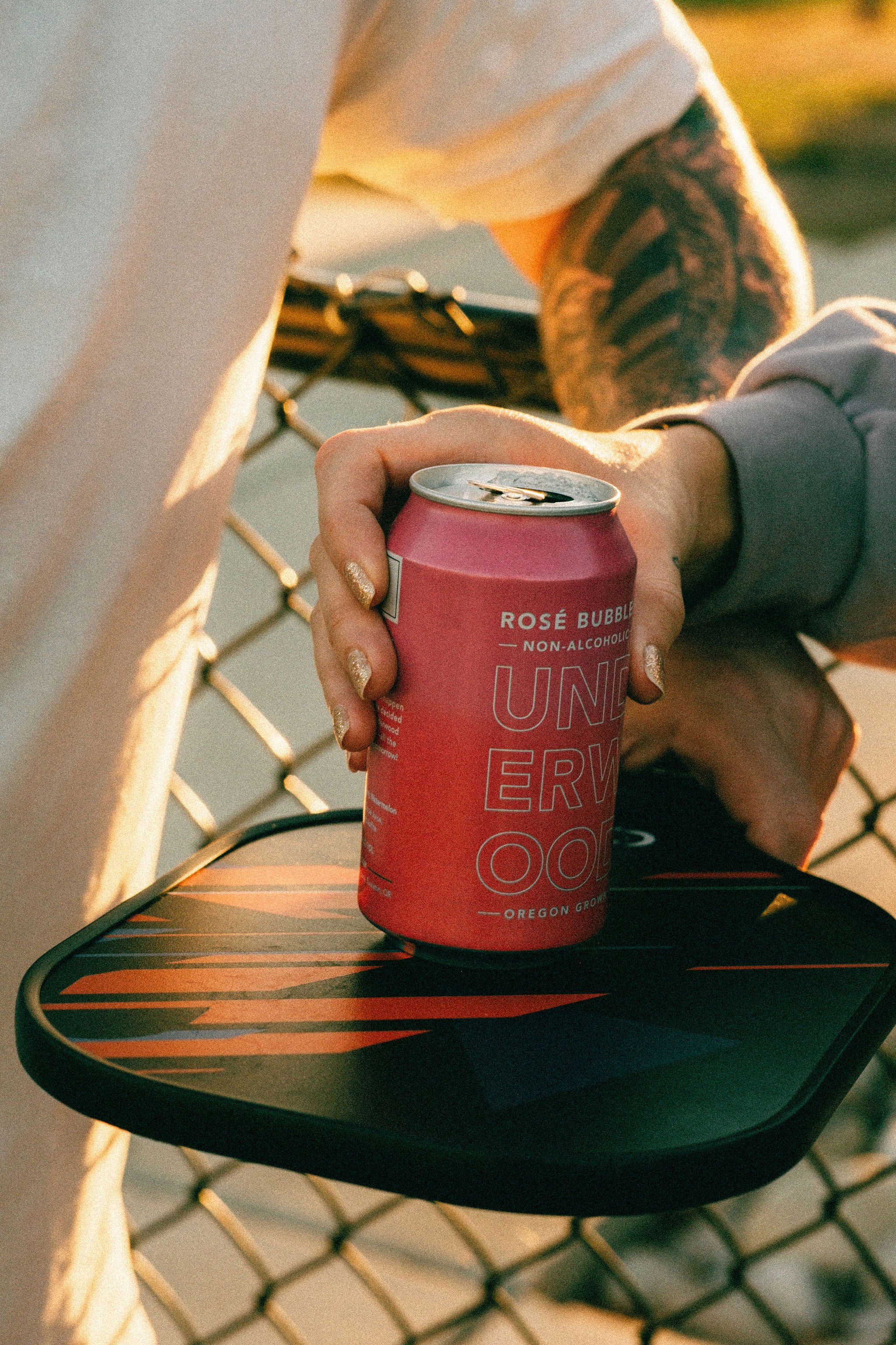Person holding a pink can of Underwood NA Rosé Bubbles on a paddleboard near a chain-link fence.