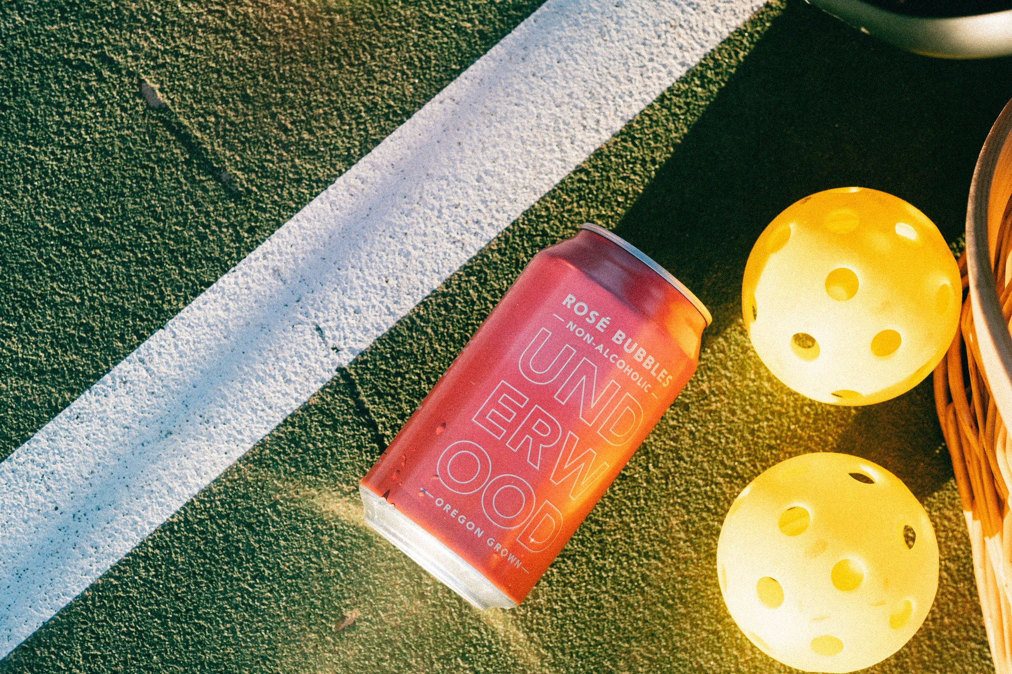 Red can of Underwood NA Rosé Bubbles on a green sports court next to two yellow perforated balls and a wicker basket.