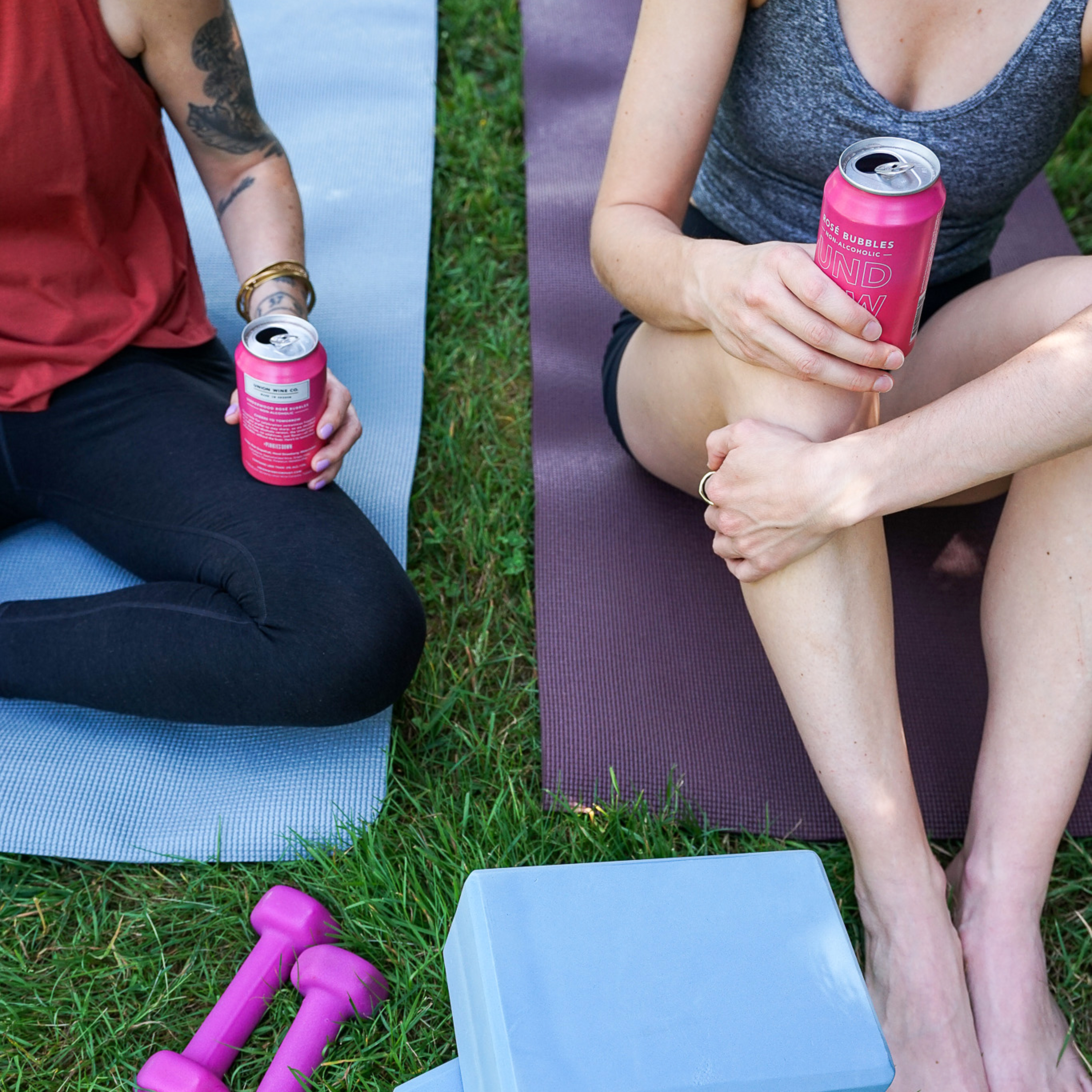 Two women sitting on yoga mats outdoors holding pink cans of Underwood NA Rosé Bubbles, with pink dumbbells and a yoga block on the grass nearby.