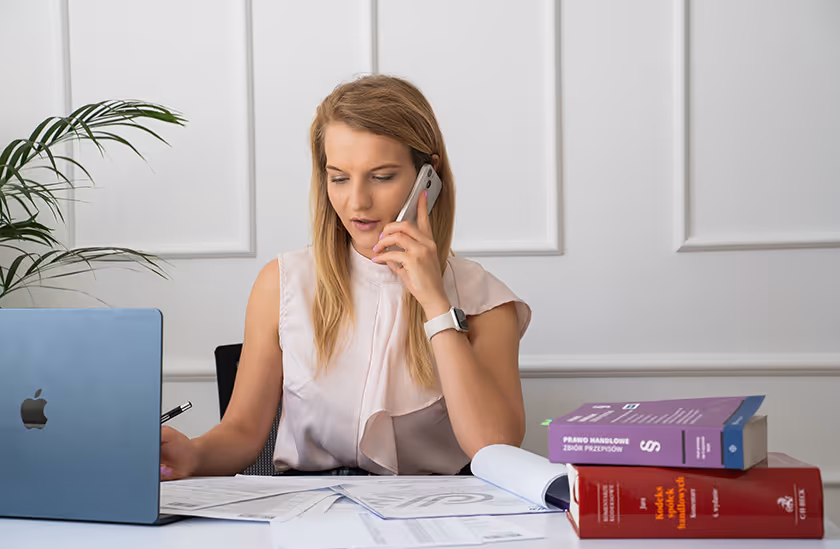 Kobieta rozmawia przez telefon przy biurku z laptopem, dokumentami i dwoma grubymi książkami prawnymi.