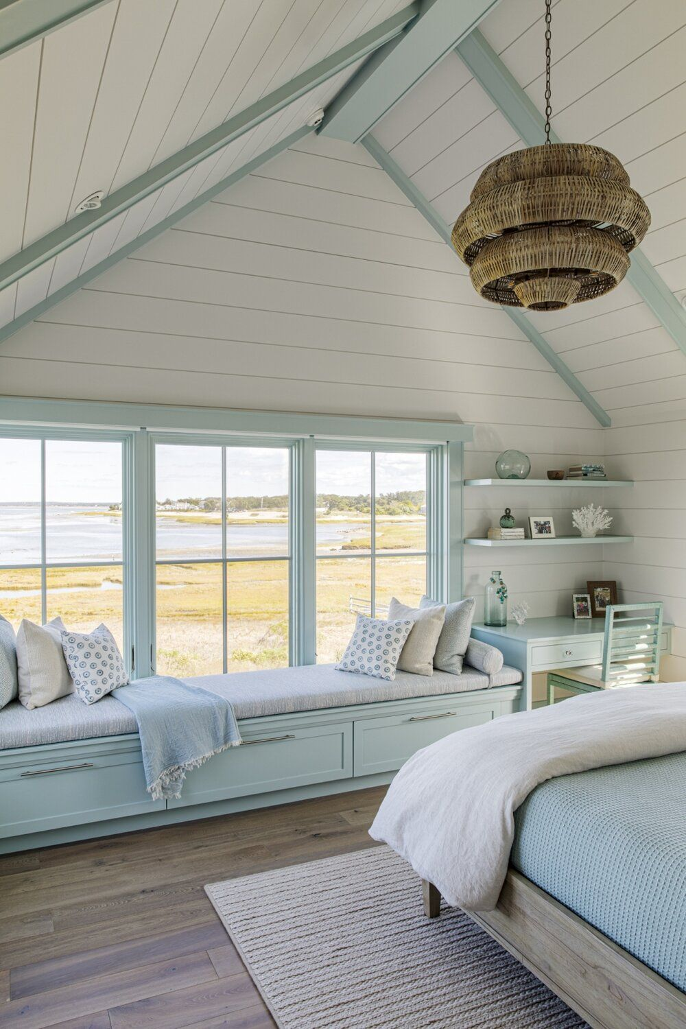 Cozy bedroom with vaulted ceiling, built-in window bench with cushions, light blue desk, and large windows overlooking a marshland.