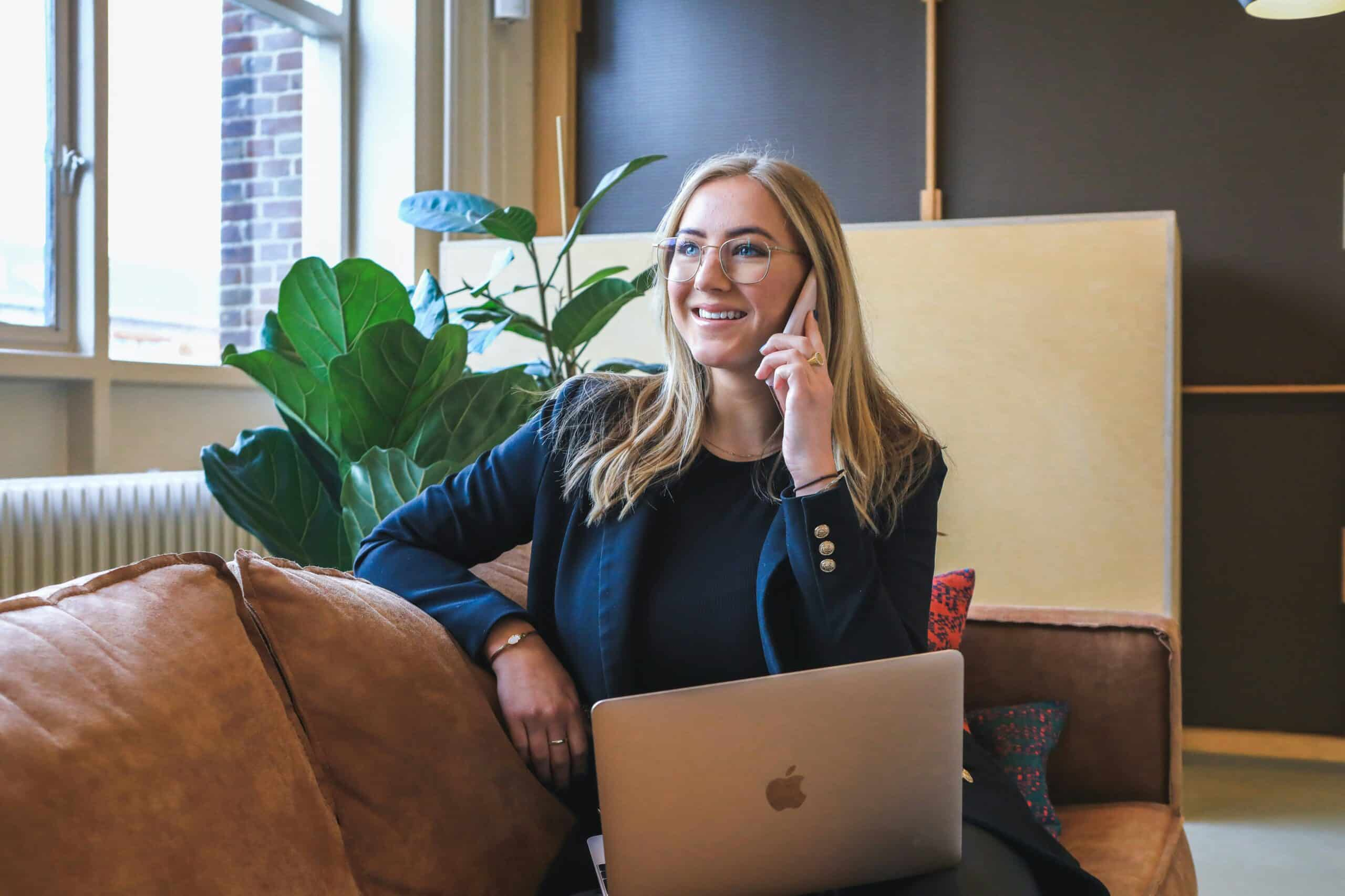 Smiling woman with glasses sitting on a brown couch with a laptop on her lap, talking on a smartphone in a cozy, plant-decorated office.