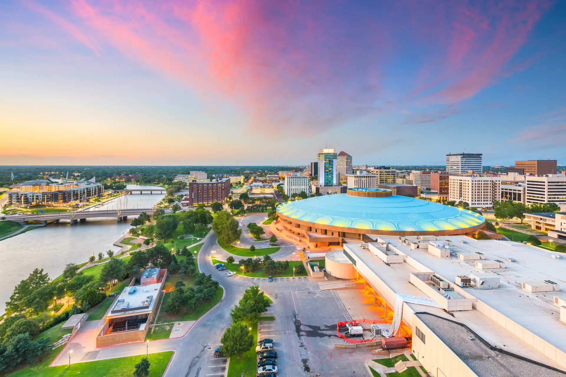 Aerial view of a city at sunset featuring a large round building with a blue roof near a river and multiple bridges.