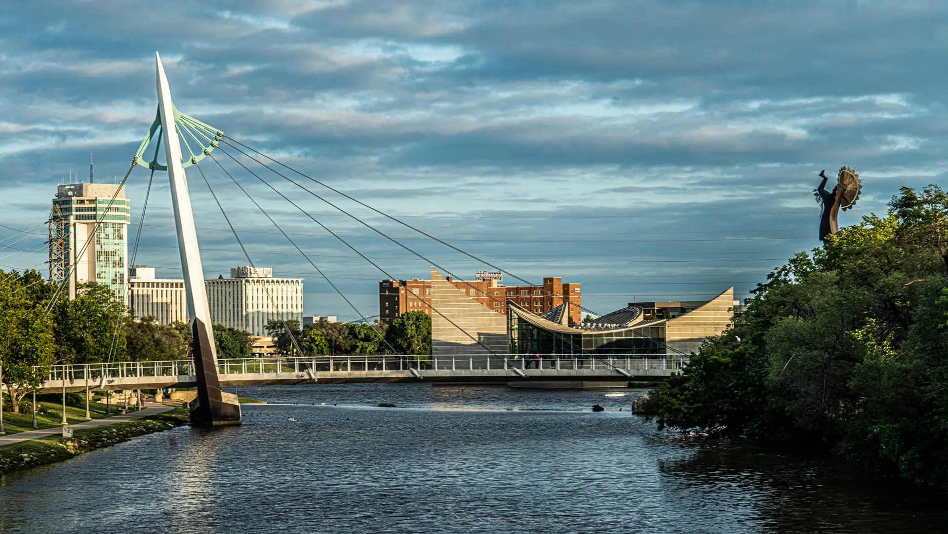 Modern cable-stayed pedestrian bridge over a river with city buildings and a statue in the background under a partly cloudy sky.