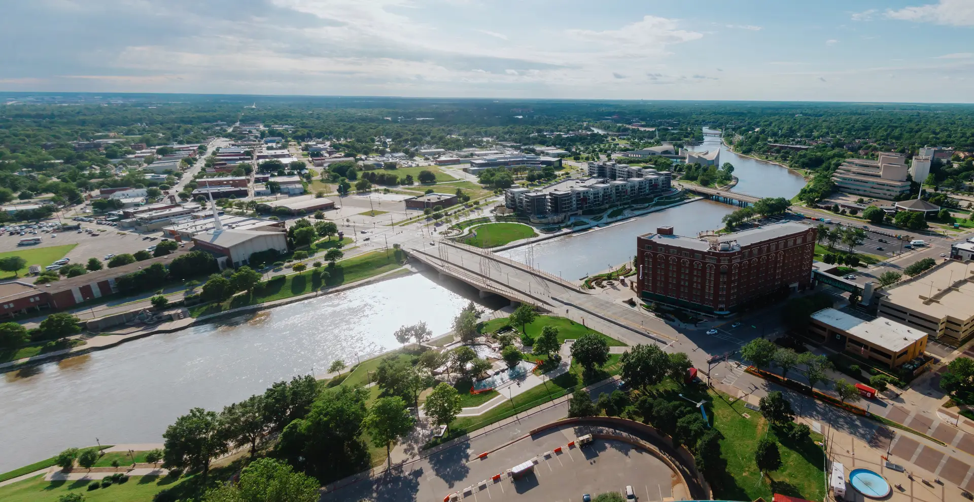 Aerial view of a city river with a bridge, parks, buildings, and green trees under a partly cloudy sky.
