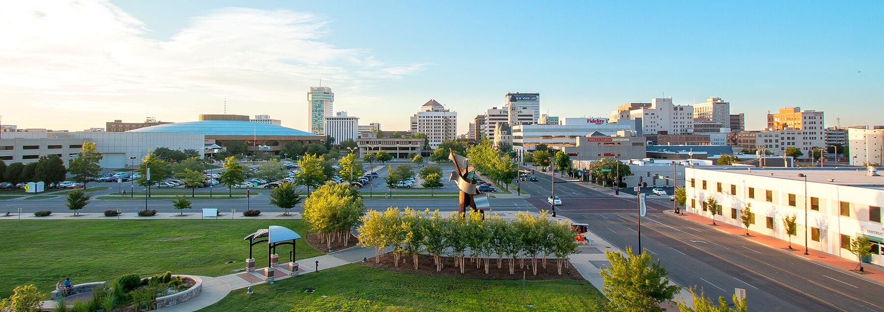 Downtown cityscape with modern buildings, a large round-roofed arena, green park area with trees, pathways, and a metal sculpture in the foreground under a clear blue sky.