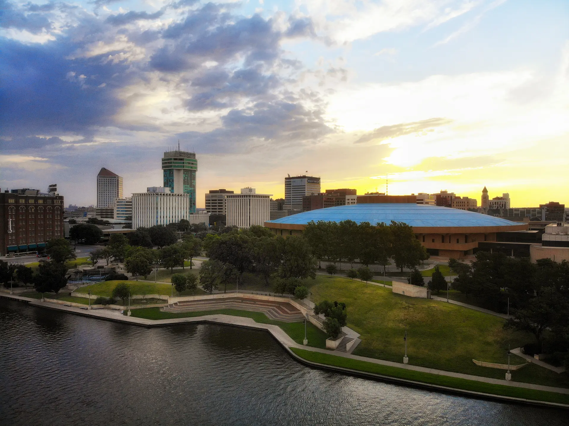 View of downtown Wichita skyline at sunset with a river and park in the foreground and a large domed building with a blue roof.