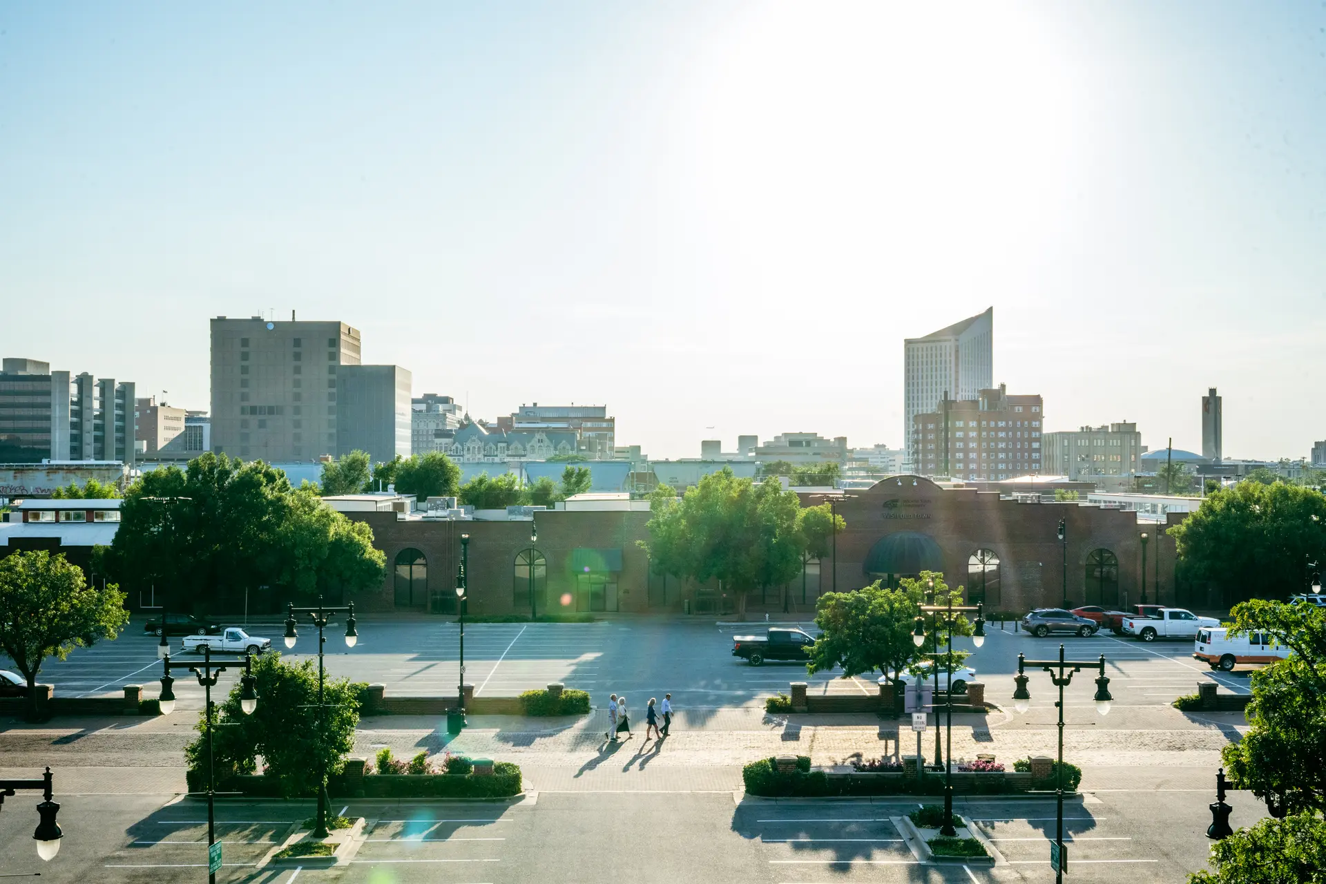 Urban street scene with a few people walking on a sidewalk, parked vehicles, green trees, and city buildings under a bright sun.