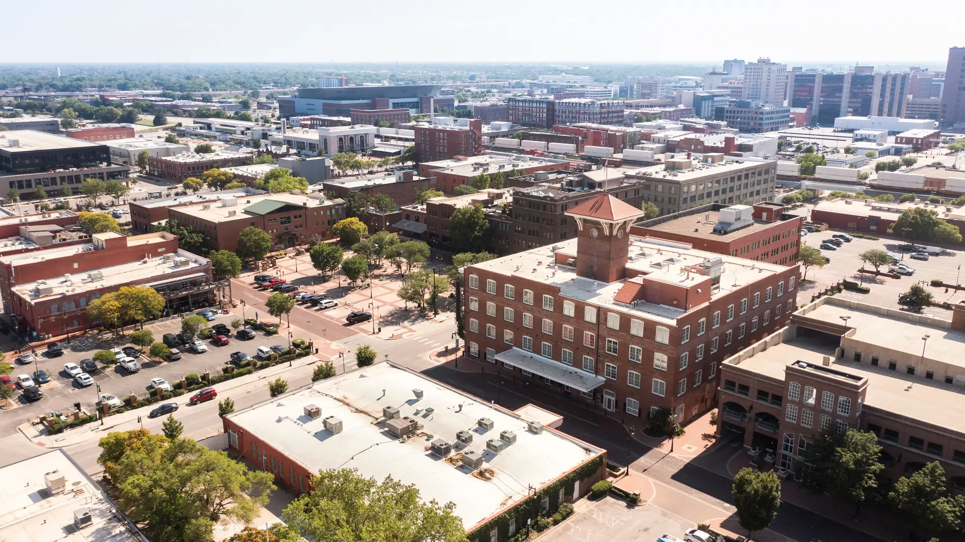 Aerial view of a city with multiple brick buildings, a clock tower, trees, and parked cars on a clear day.