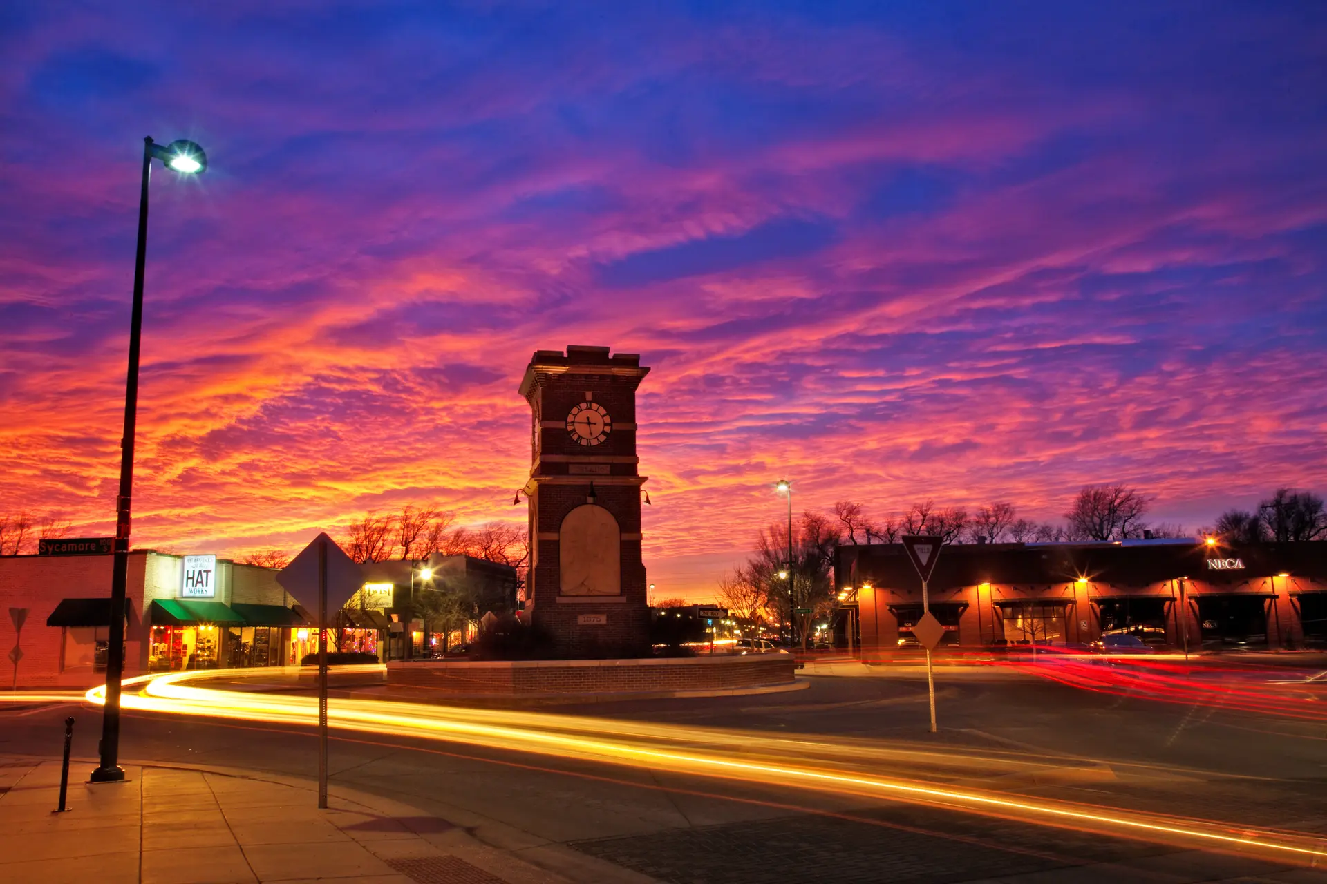 Sunset sky with vibrant orange and purple clouds above a town square featuring a brick clock tower and light trails from passing cars.