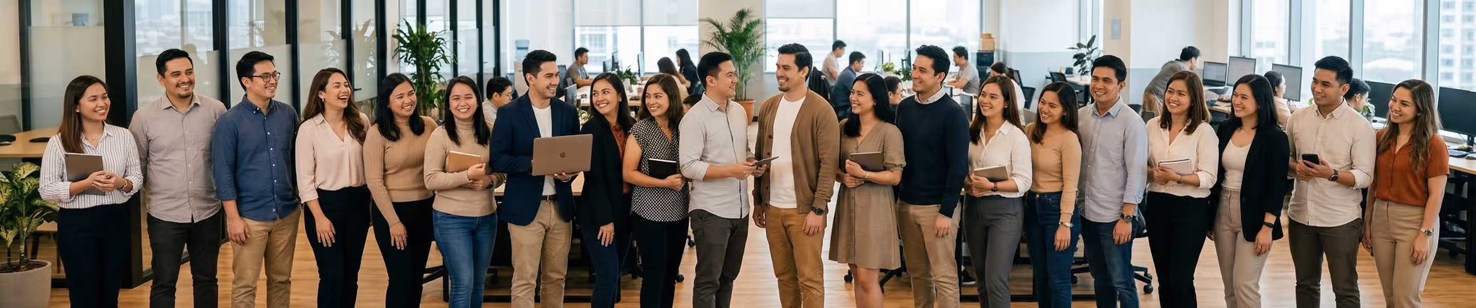 Group of diverse young professionals standing and smiling in a modern office with desks and computers in the background.