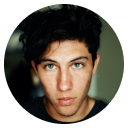 Young man with dark curly hair and blue eyes looking directly at the camera against a blurred background.