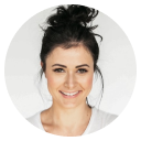 Smiling woman with dark hair in a messy bun wearing a white shirt against a light gray background.