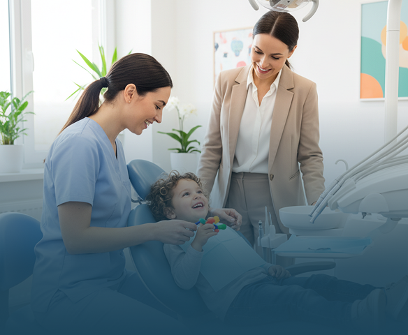 A young boy with curly hair smiles while sitting in a dental chair as a female dentist in blue scrubs and a woman in a beige suit stand nearby, all in a bright dental office with plants and colorful artwork.