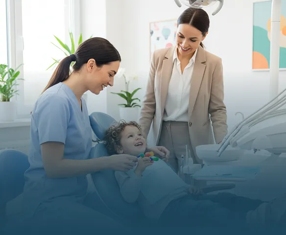 A young boy with curly hair smiles while sitting in a dental chair as a female dentist in blue scrubs and a woman in a beige suit stand nearby, all in a bright dental office with plants and colorful artwork.