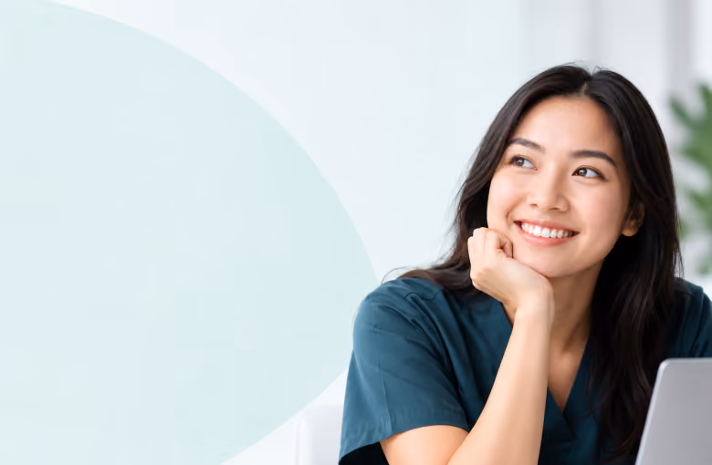 Smiling young woman with long dark hair wearing a teal shirt, resting her chin on her hand and looking to the side, seated at a desk with a laptop.