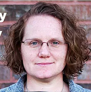 Headshot of a Carrie Williamson with short curly brown hair, glasses, and a neutral expression, standing in front of a brick wall.